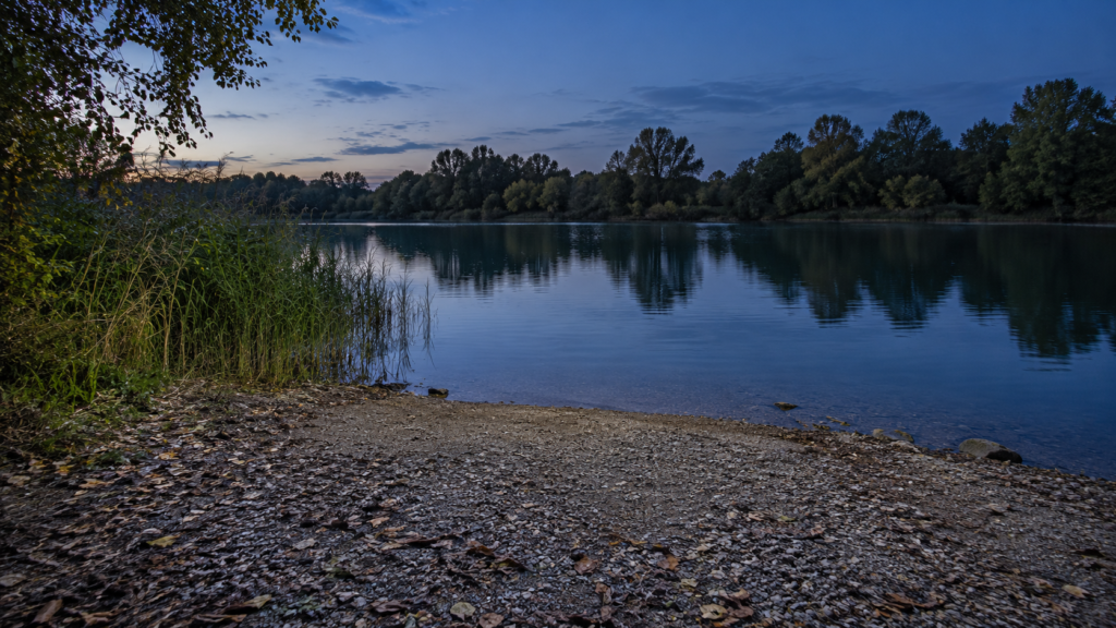 Breiter kiesiger Uferzugang an einem See in der Abenddämmerung mit Pflanzen am linken Rand