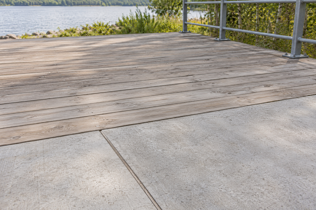 Detailaufnahme eines Übergangs von hellen Betonplatten auf eine freie Holzplattform mit Metallgeländer am Rand.