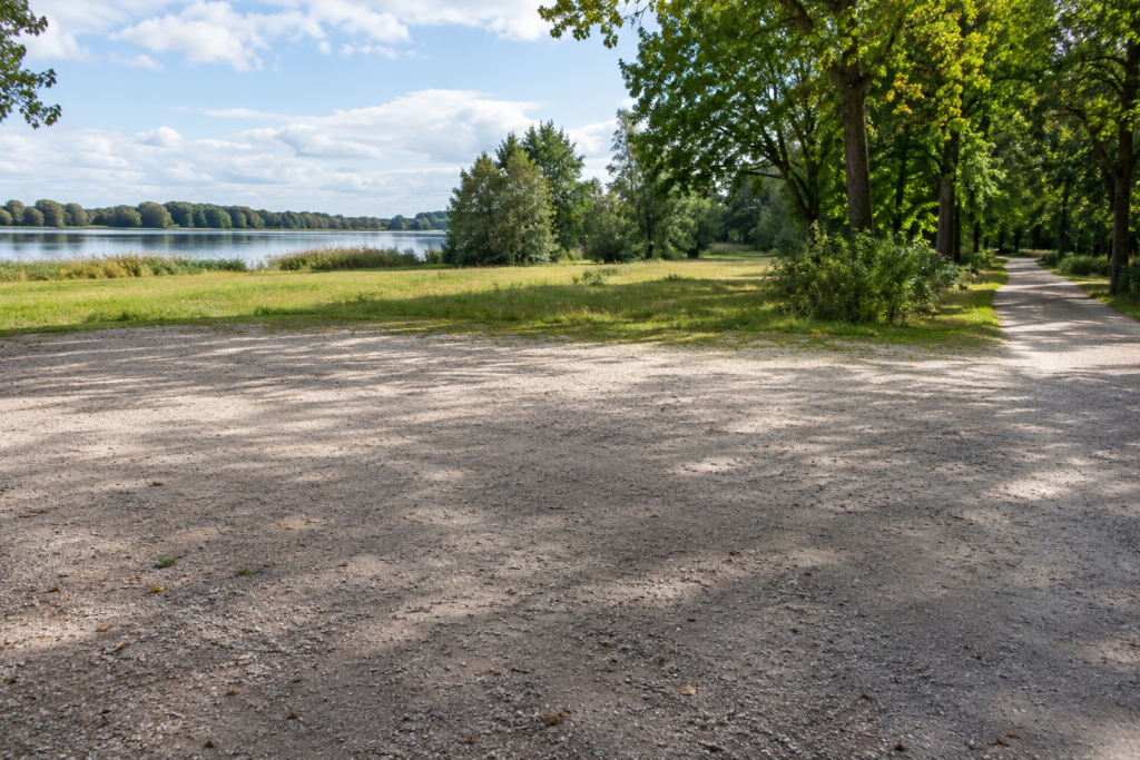 Breite freie Kiesfläche mit rechts anschließendem Weg; zwischen der Fläche und dem See liegt eine ausgedehnte Wiese.