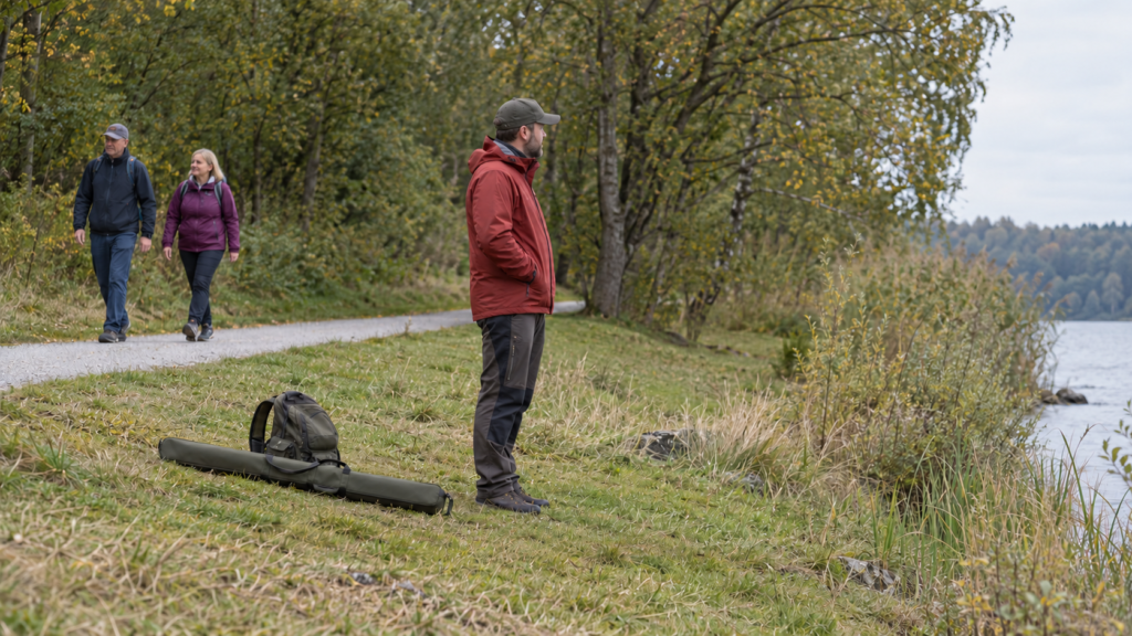 Eine Person steht neben geschlossenem Rutenfutteral und Rucksack auf der Uferwiese; dahinter gehen zwei Menschen auf einem Kiesweg.