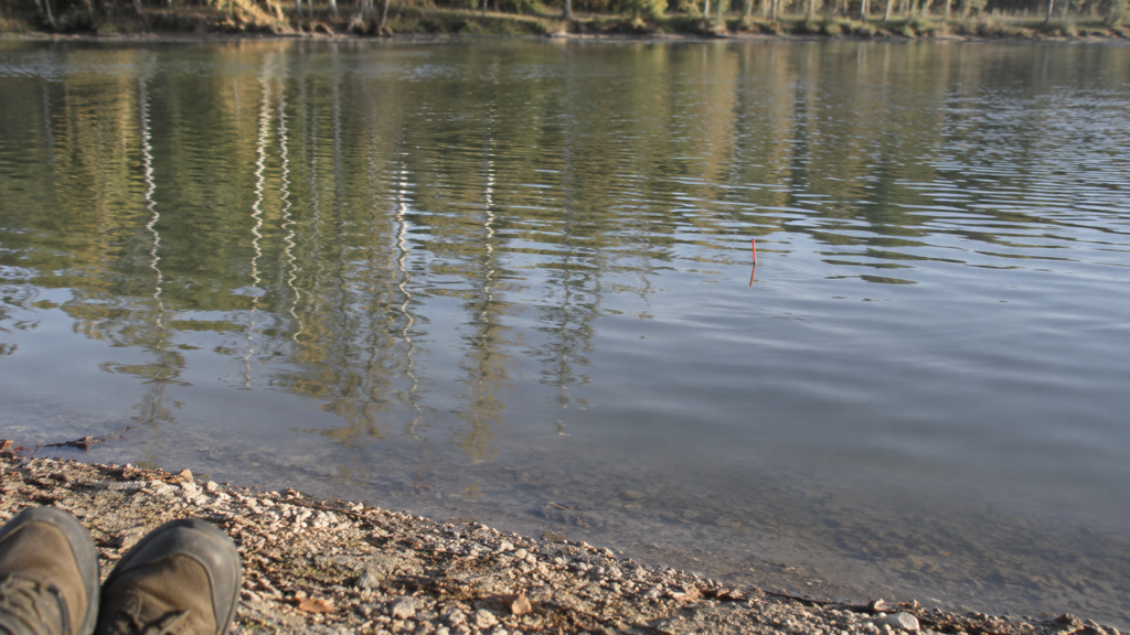 Vom trockenen Kiesrand aus ist eine rote Posenantenne auf dem Wasser zu sehen; unten erscheinen die Spitzen zweier Wanderschuhe.