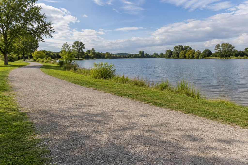 Breiter trockener Kiesweg mit freien Standflächen und einem Grasstreifen neben einem See