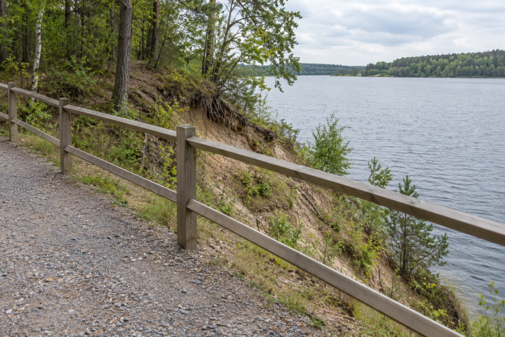 Befestigter Kiesweg mit Holzgeländer vor einer steilen erodierten Uferböschung am See